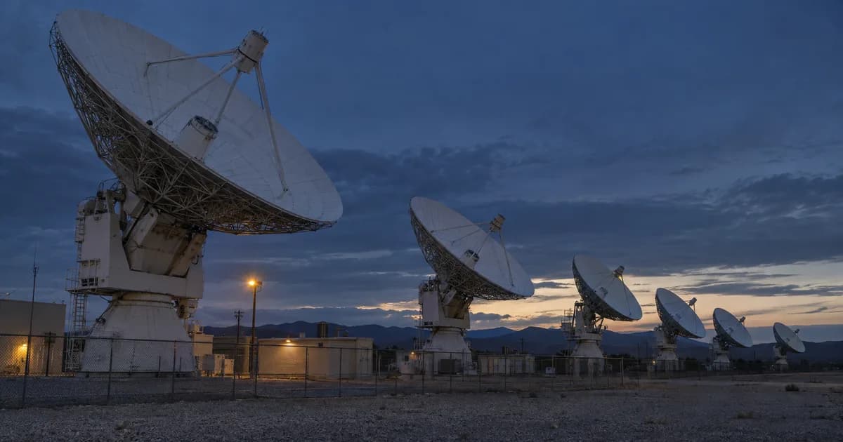A row of large white satellite uplink dishes angled skyward at dusk on a gravel compound behind a chain-link fence, mountains on the horizon.