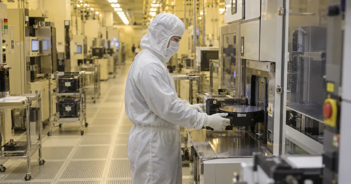 A technician in a white cleanroom bunny suit and face mask loading a silicon wafer carrier into a processing tool inside a semiconductor fabrication plant, with rows of equipment receding down the aisle under yellow photolithography lighting.