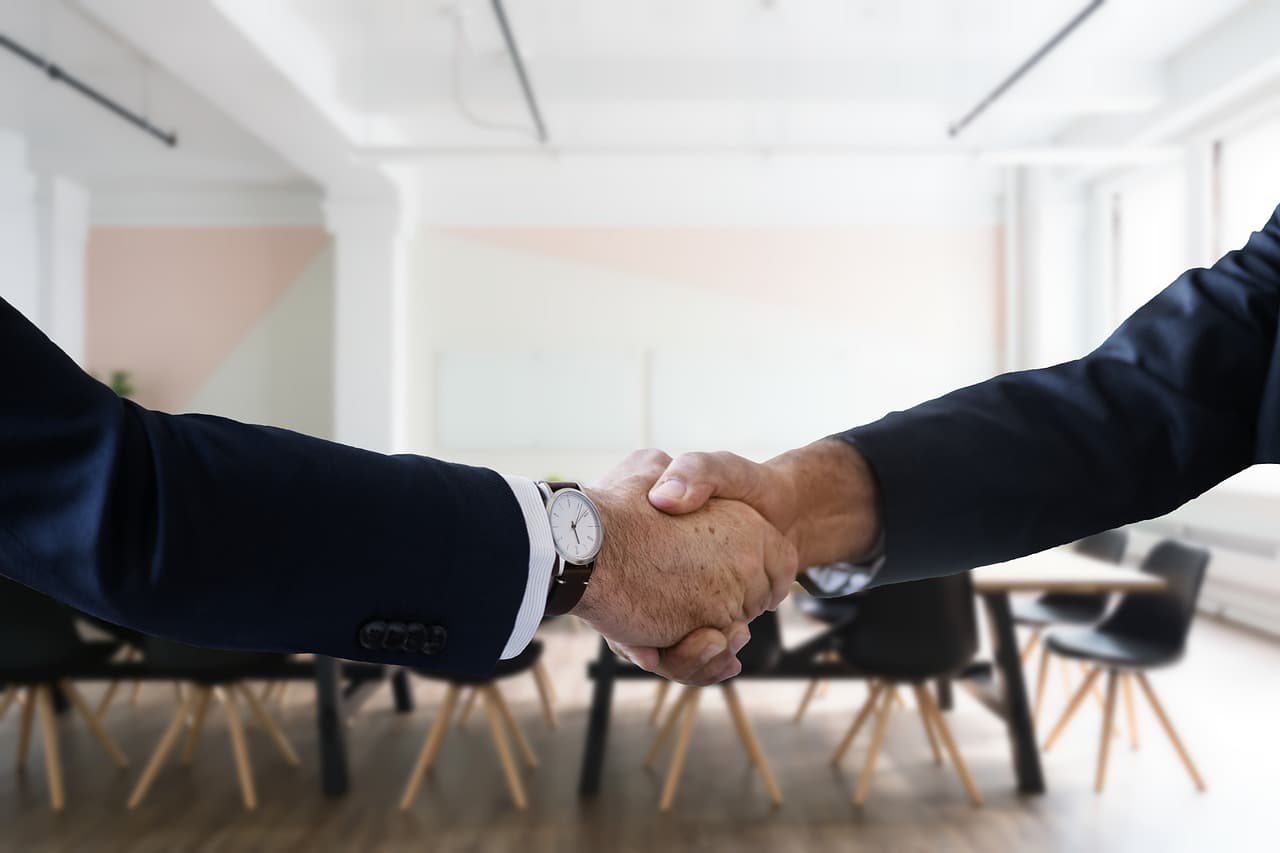 Two people in business suits shaking hands across a table in a bright, largely empty office, representing hiring in a slow-growth labor market
